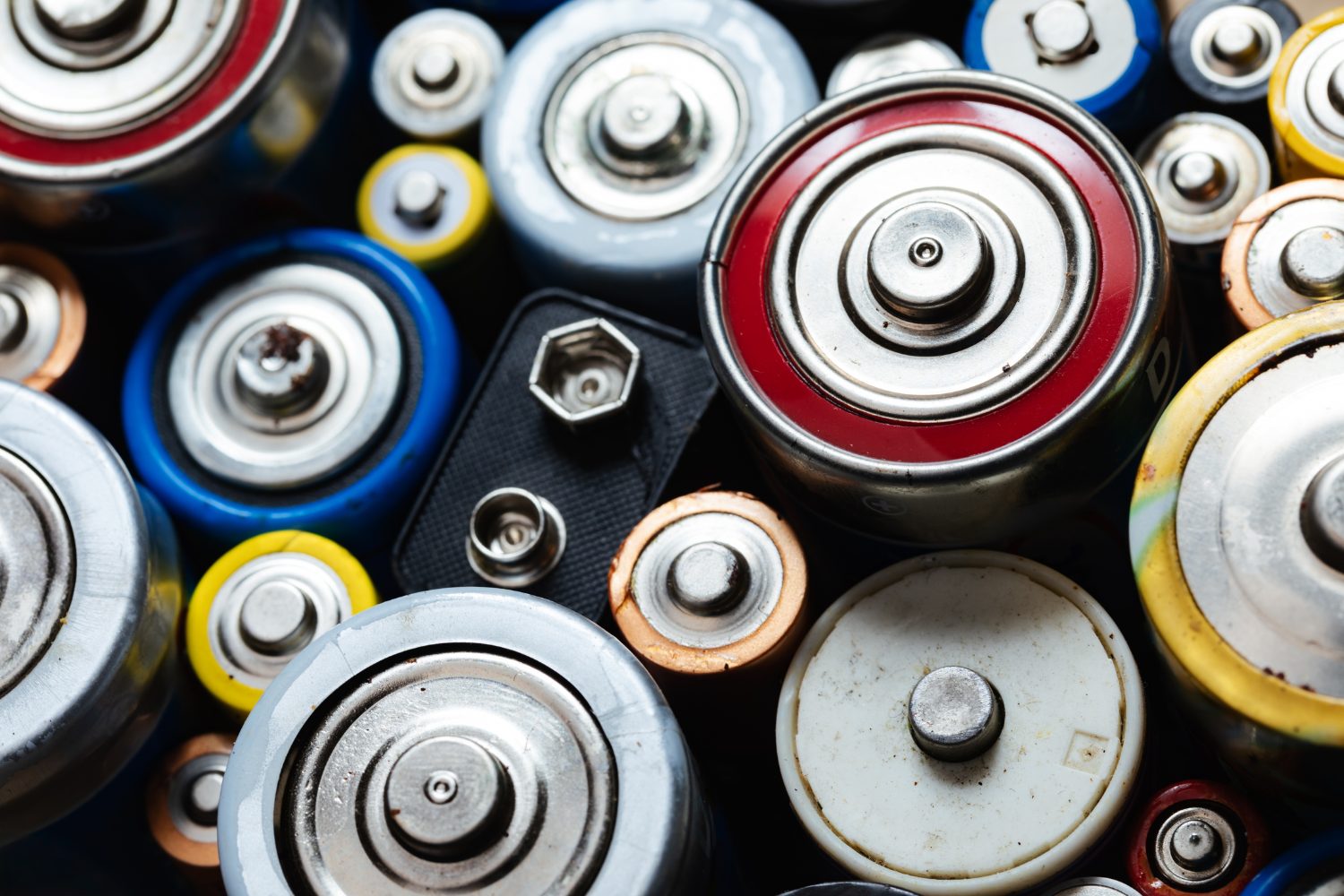Close-up of assorted household batteries arranged tightly, showing the positive terminals of D, C, AA and AAA cells in various colors alongside a 9V connector.