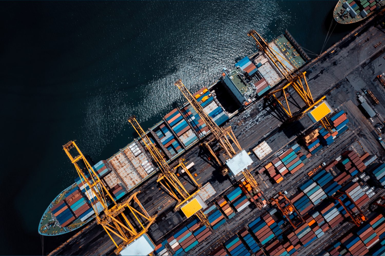 Aerial view of a busy container port with four large yellow gantry cranes along the dock loading and unloading multicolored shipping containers from cargo ships moored beside dark, reflective water.