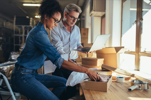 Two colleagues in a sunlit packing area preparing orders — woman placing sweater into a box while man checks a laptop
