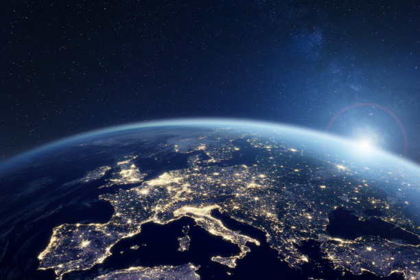 Satellite view of Earth at night focusing on Europe aglow with city lights, the planet's curved horizon and atmospheric glow, and a bright sunrise emerging against a star-filled sky.