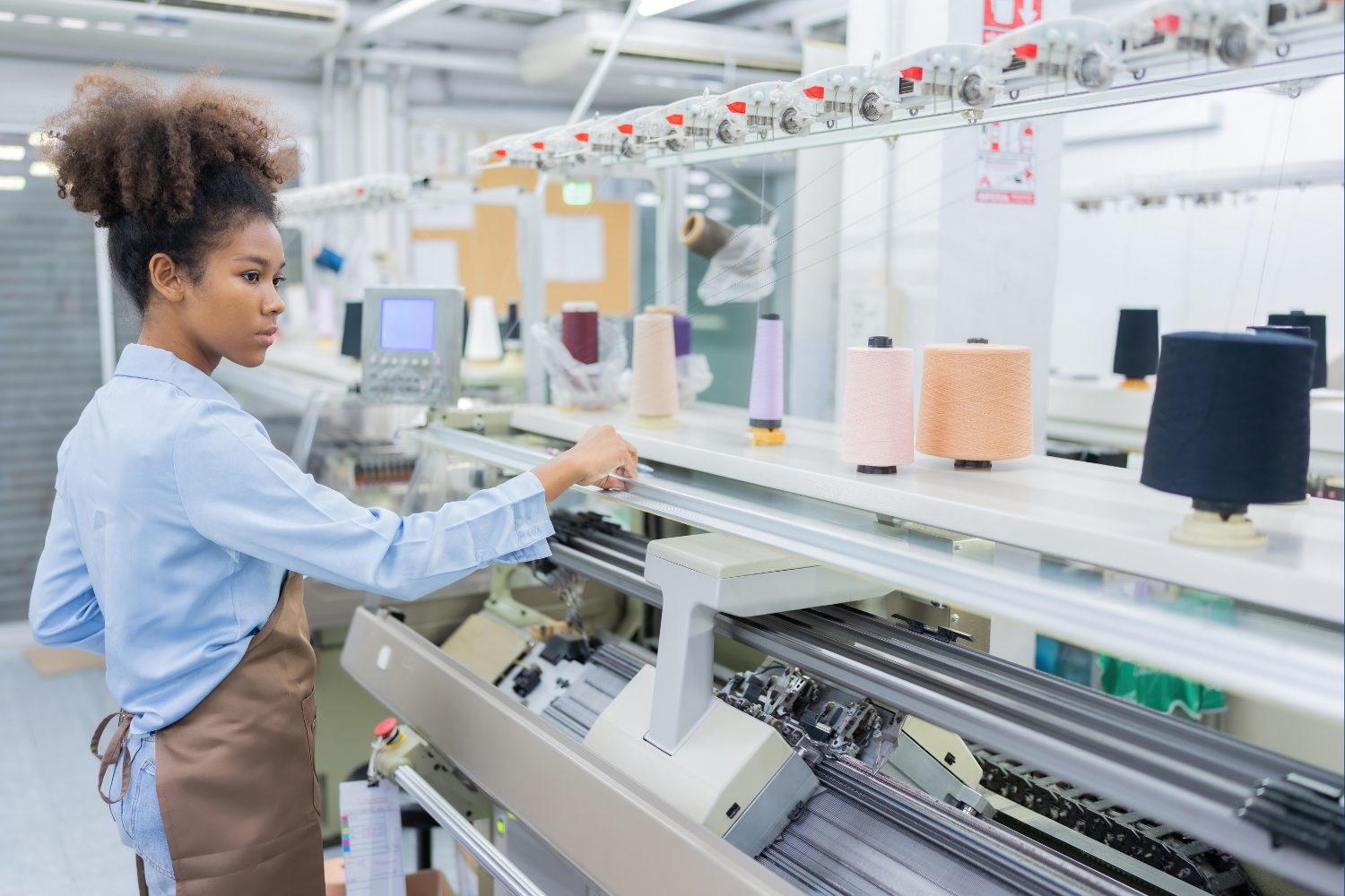 Young woman with curly hair in a high puff, wearing a light blue shirt and brown apron, adjusts a large industrial knitting machine while pastel and dark thread spools line the machine in a bright factory workspace.