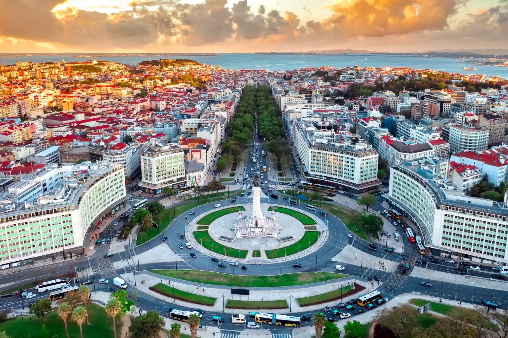 Aerial view of a bustling roundabout with a central monument, surrounded by an array of buildings at sunset, with the sea and horizon in the background.