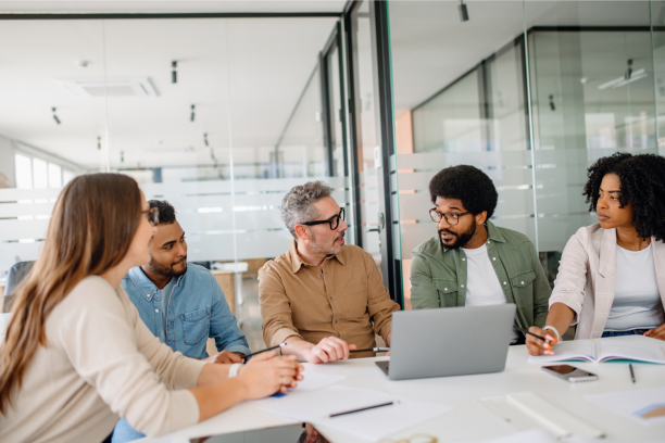 Five colleagues of varying ages seated around a conference table in a glass-walled office, engaged in discussion while reviewing a laptop, notebooks and papers.