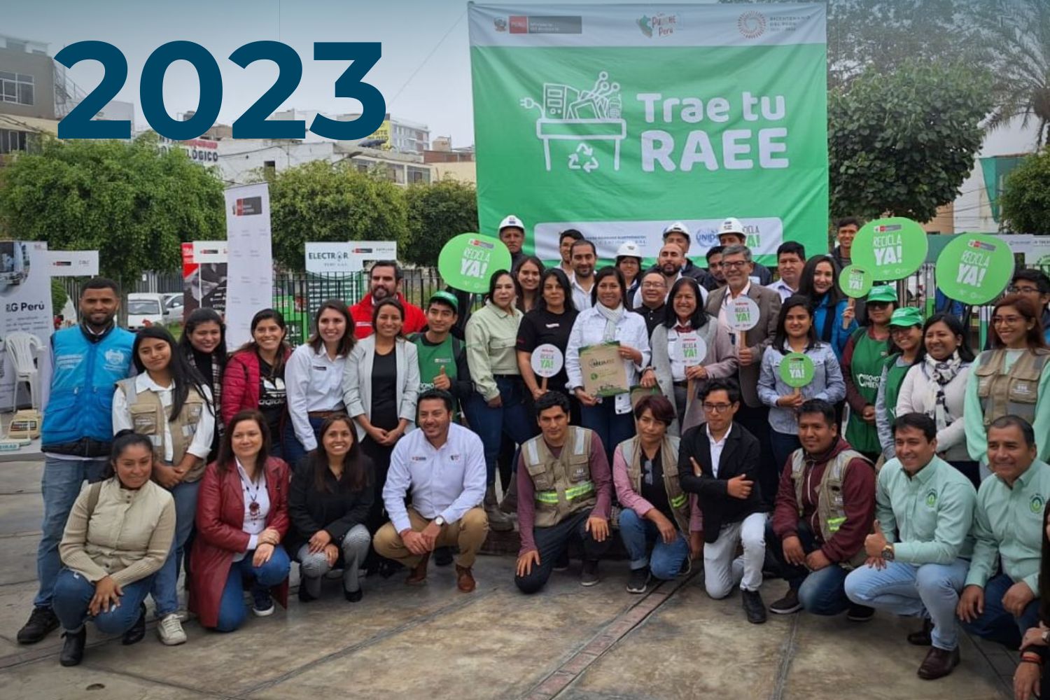 Un grupo de personas posa para una foto en un evento de reciclaje con una pancarta grande que dice "Trae tu RAEE" y carteles que promueven el reciclaje, con el número 2023 superpuesto en la imagen.