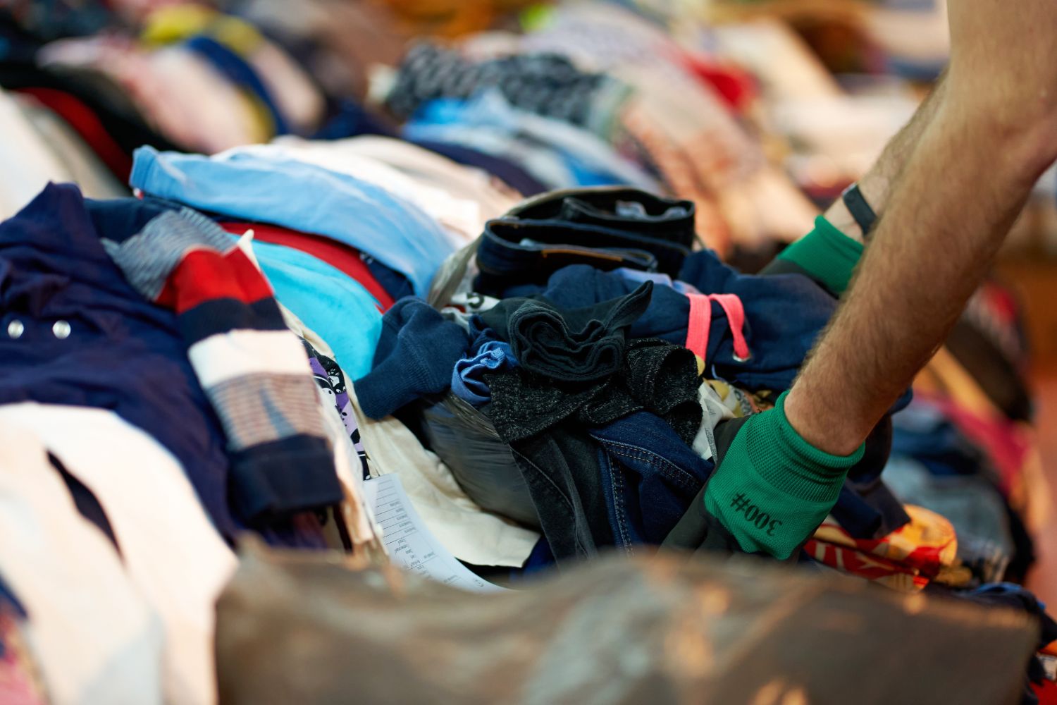 Braccio con guanti verdi che sistema una pila di vestiti colorati e piegati su un tavolo, tra magliette, jeans e calzini.