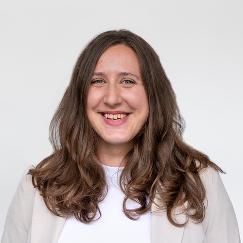 Smiling woman with long wavy brown hair wearing a light blazer over a white top against a plain light background.