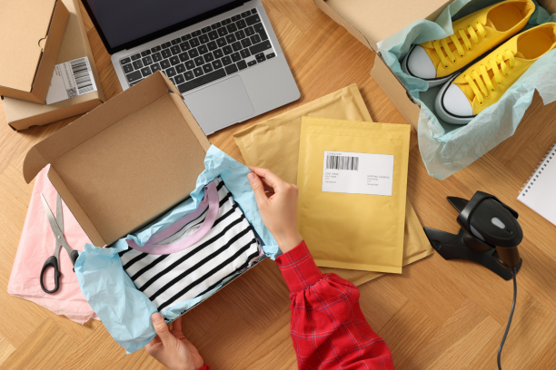 Hands in a red plaid shirt packing a black-and-white striped T‑shirt wrapped in blue tissue into a cardboard shipping box on a wooden desk surrounded by a laptop, padded mailers, yellow sneakers in a shoebox, scissors and a barcode scanner.