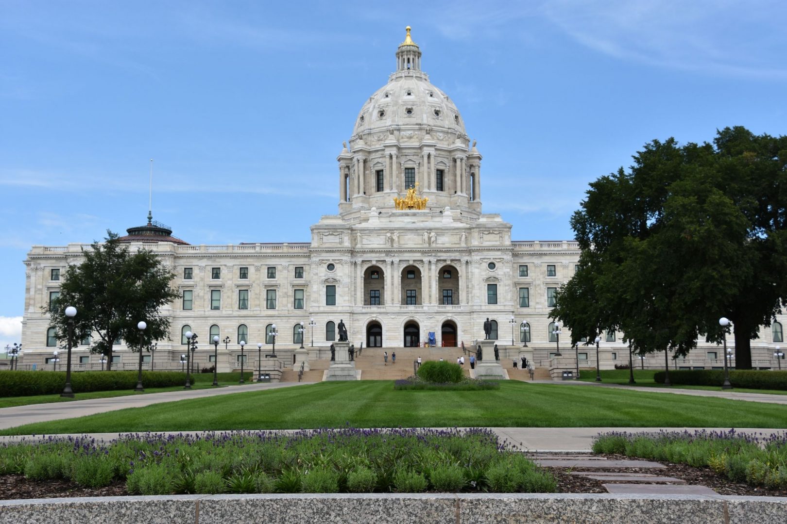 Alt text: The Minnesota State Capitol building with a central dome and gilded quadriga, flanked by green lawn and garden beds, under a partly cloudy sky.
