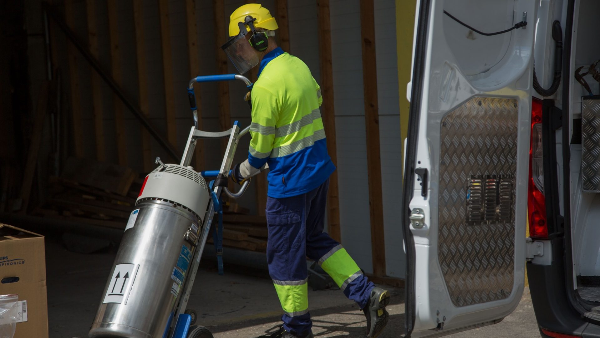 A worker in high-visibility clothing is unloading a large cylindrical tank from the back of a van using a hand trolley.