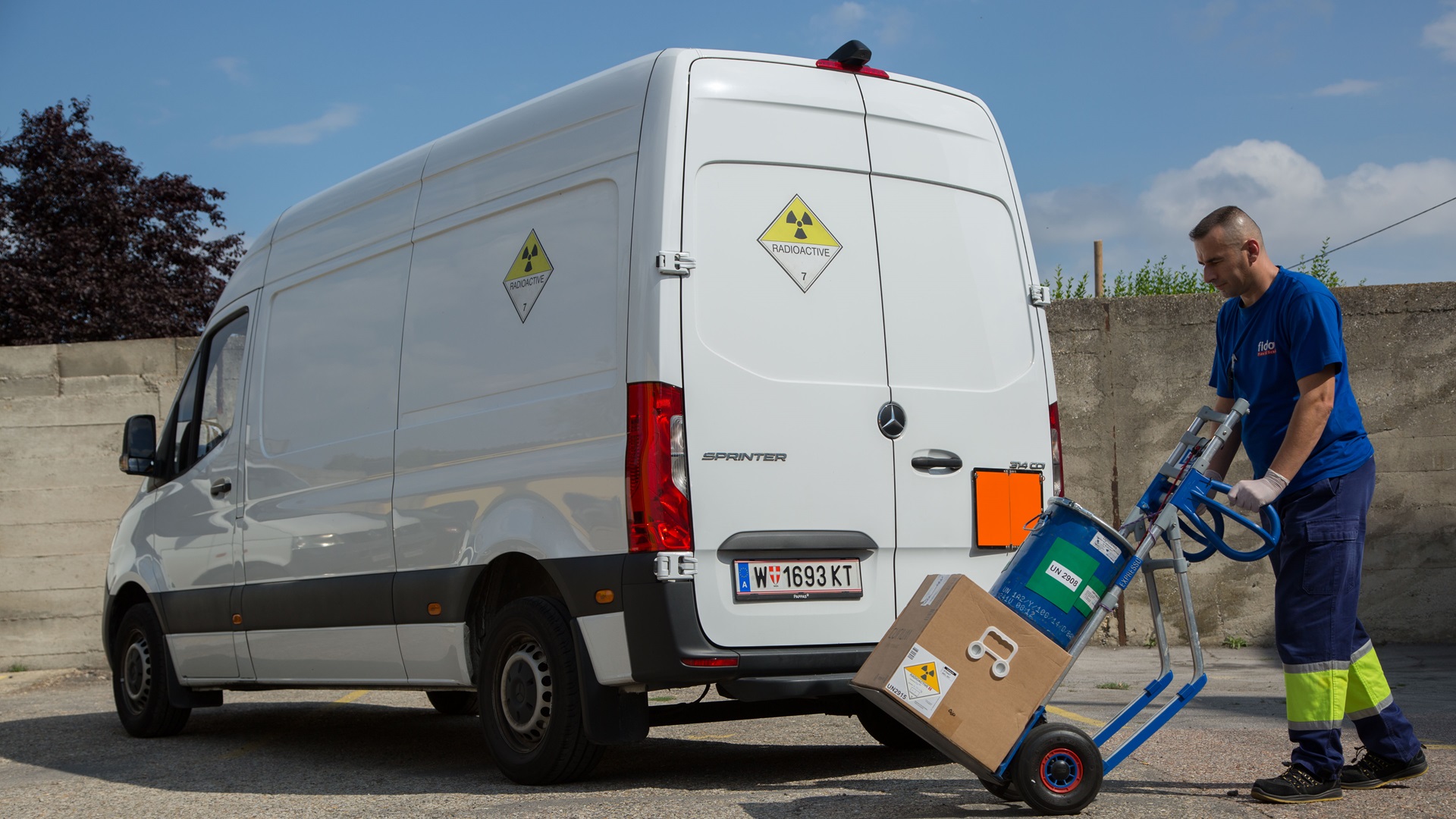 A worker in a blue uniform is using a hand truck to transport a cardboard box with hazardous material labels next to a white Sprinter van marked with radioactive warning signs.