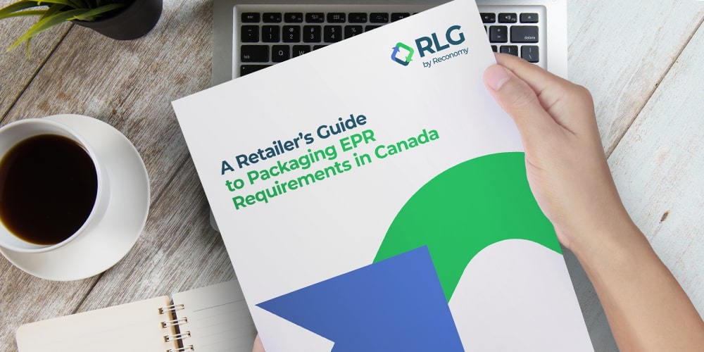 Hands holding a brochure titled "A Retailer's Guide to Packaging EPR Requirements in Canada" featuring the RLG logo, placed over a laptop on a light wooden desk next to a cup of coffee and an open notebook.