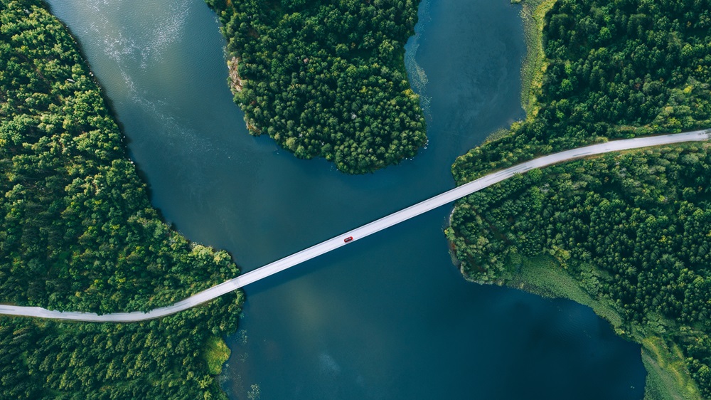 Luftfoto af en lang, smal bro over en blå sø omgivet af tætte grønne skove, med en lille rød bil på broen.