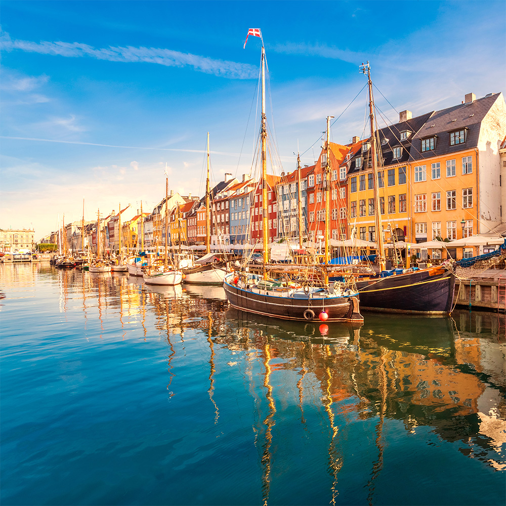 Colorful buildings line a canal with several boats moored along the quayside, reflecting onto the water's surface under a clear sky.