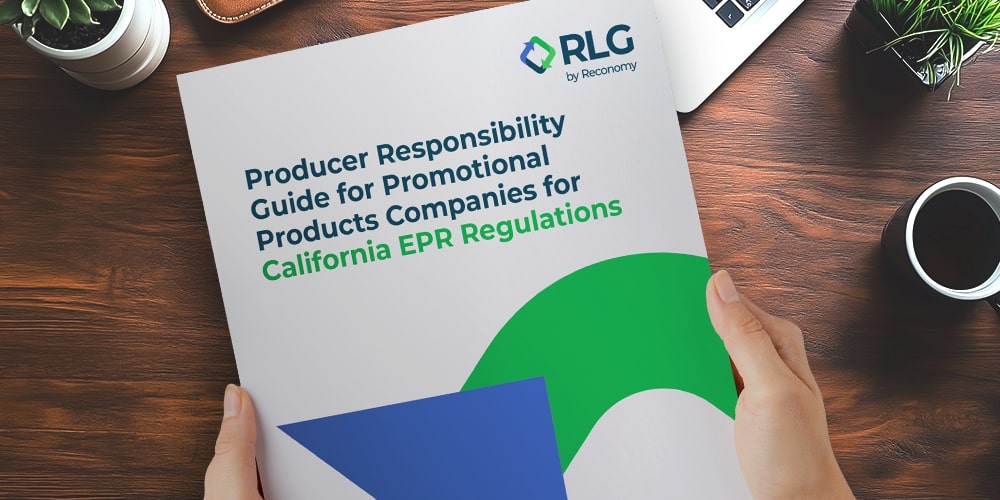 Hands hold a printed guide titled Producer Responsibility Guide for Promotional Products Companies for California EPR Regulations, featuring blue and green graphics, on a wooden desk beside a laptop, coffee cup, and potted plant.