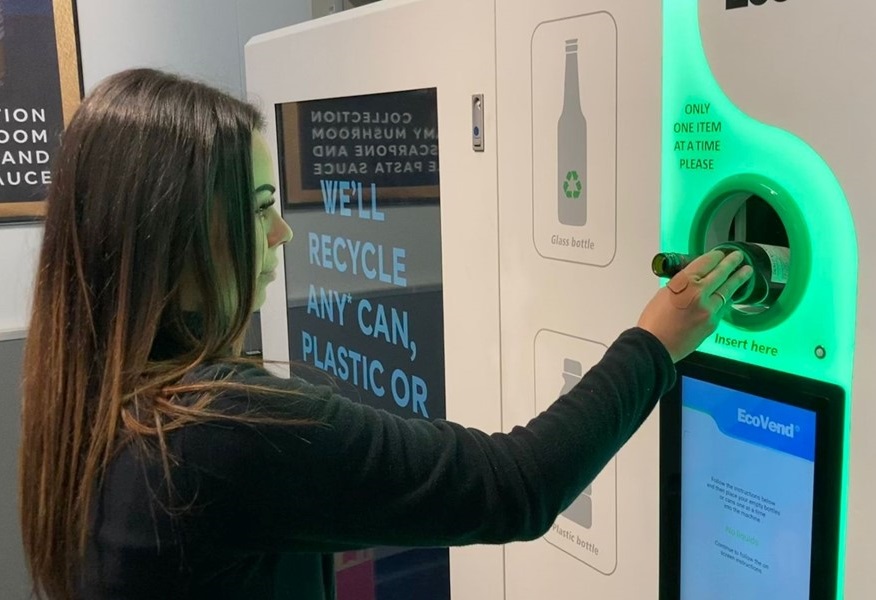 A person is recycling a bottle at an 'EcoVend' reverse vending machine, which emphasizes recycling cans, plastic, and glass bottles.