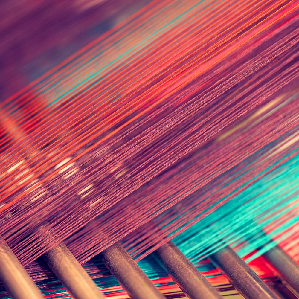 Close-up of colorful thread strands on a loom, with a focus on red and teal hues, creating an abstract textile pattern.