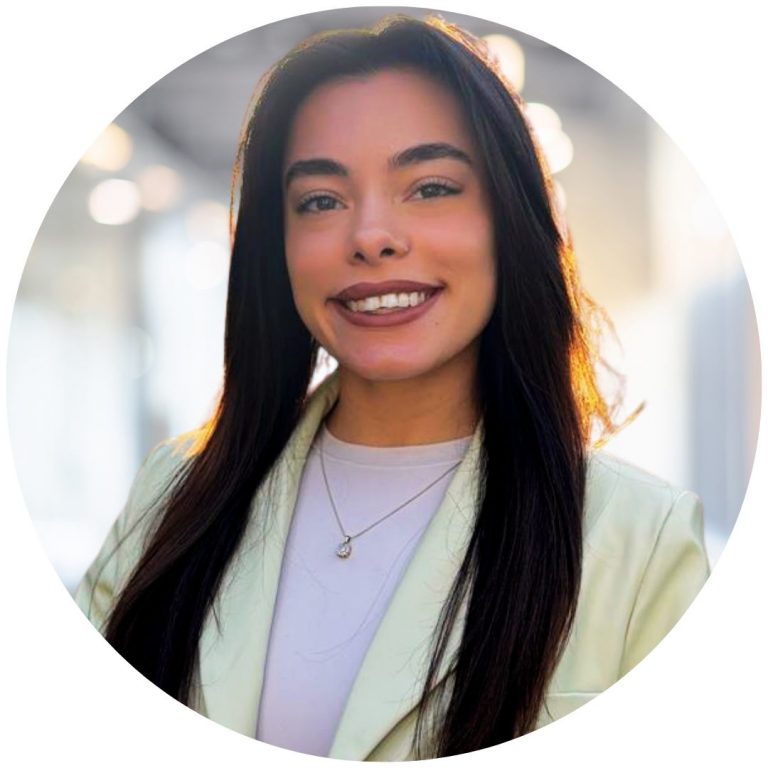 Smiling young woman with long dark hair wearing a light-colored blazer and a pendant necklace, photographed in a warm backlit indoor setting with a blurred bokeh background.