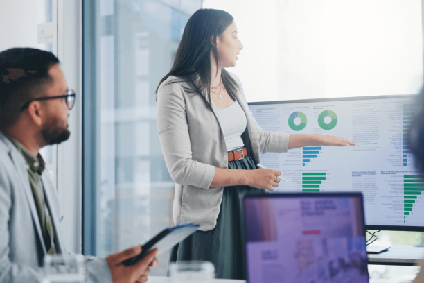 Woman in a gray blazer presenting in a glass-walled office, pointing at a monitor showing green pie and bar charts while a seated colleague with glasses holds a tablet and a laptop screen blurs in the foreground.