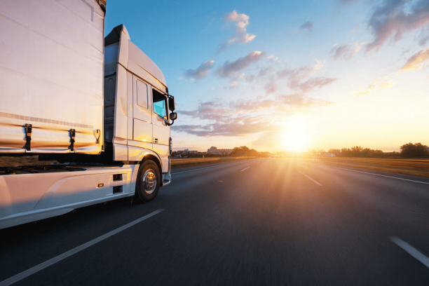 White semi-truck cab and trailer captured from the left side driving on a multi-lane highway toward a bright setting sun, with motion-blurred road and golden light reflecting off the truck under a partly cloudy sky.