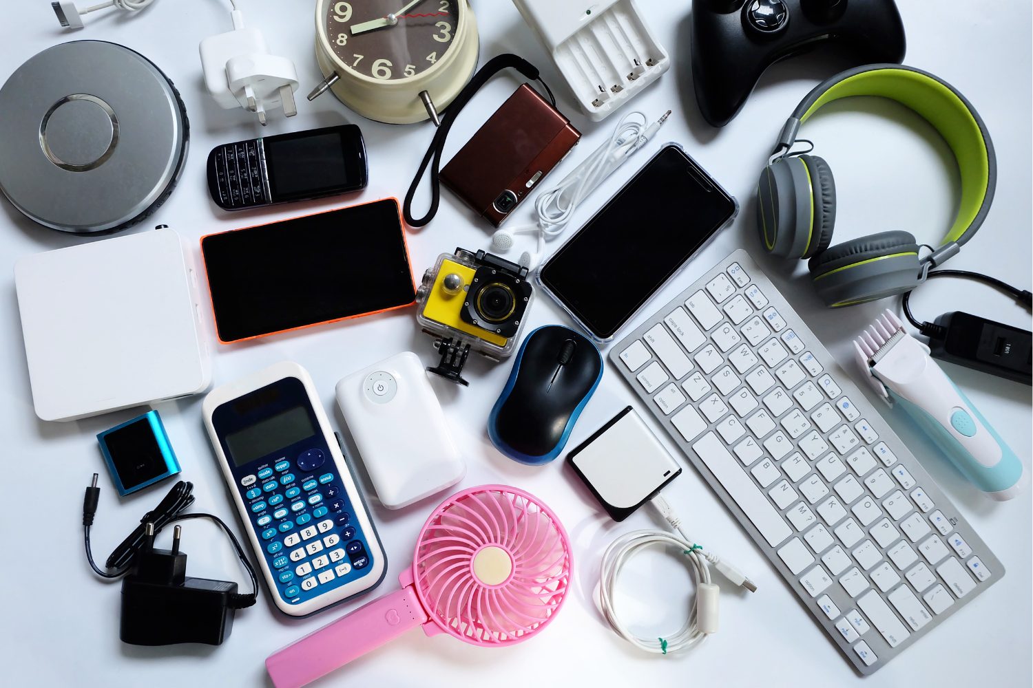 Various electronic gadgets and accessories on a white surface including a white keyboard, gray and green headphones, a black and blue computer mouse, a pink handheld fan, a calculator, a retro mobile phone, an alarm clock, a game controller, a camera, earphones, charging cables, an electric hair clipper, and a few adapters and chargers.