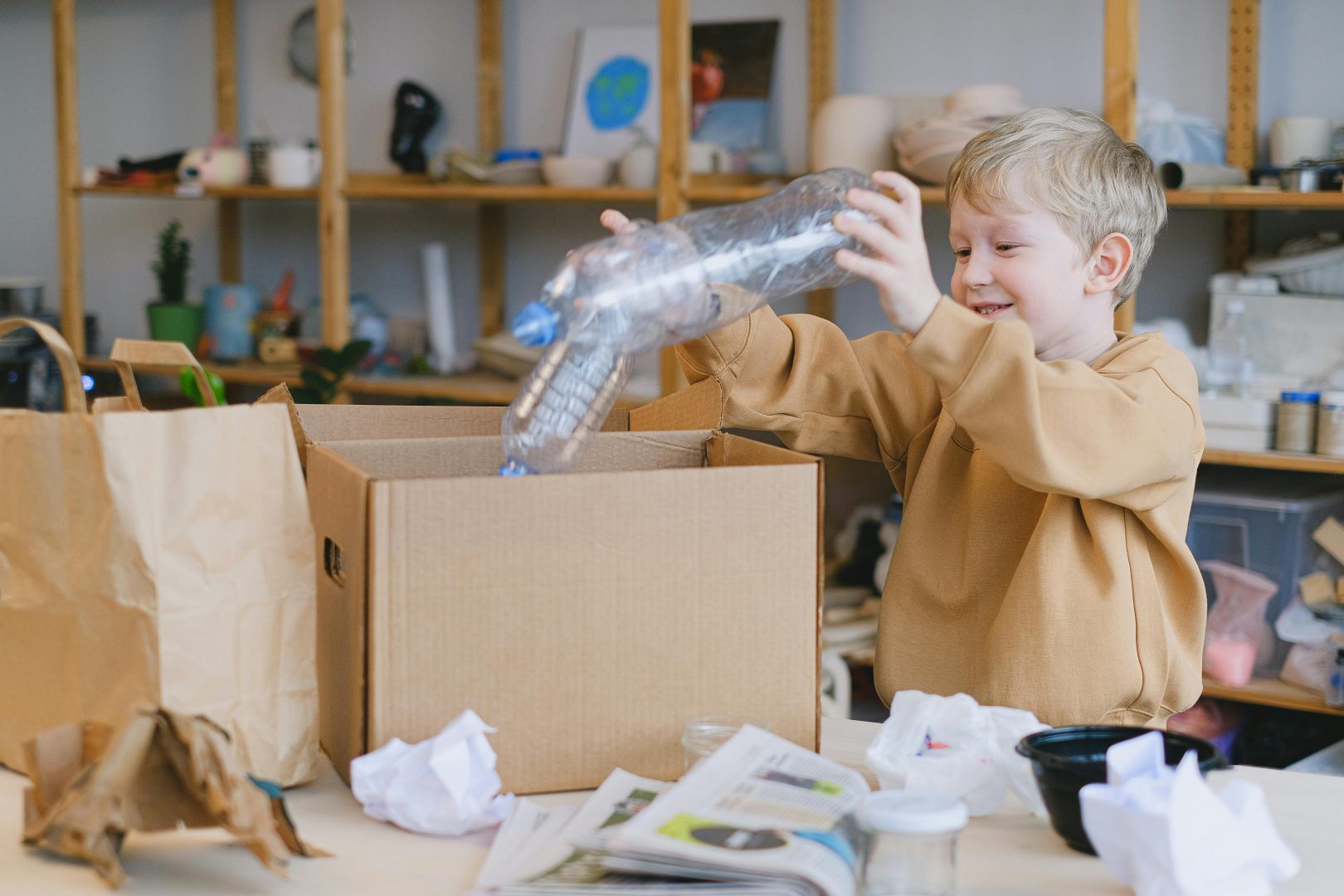 A young boy in a beige hoodie smiling as he takes a large plastic bottle out of a cardboard box in a room with shelves and various items scattered around.