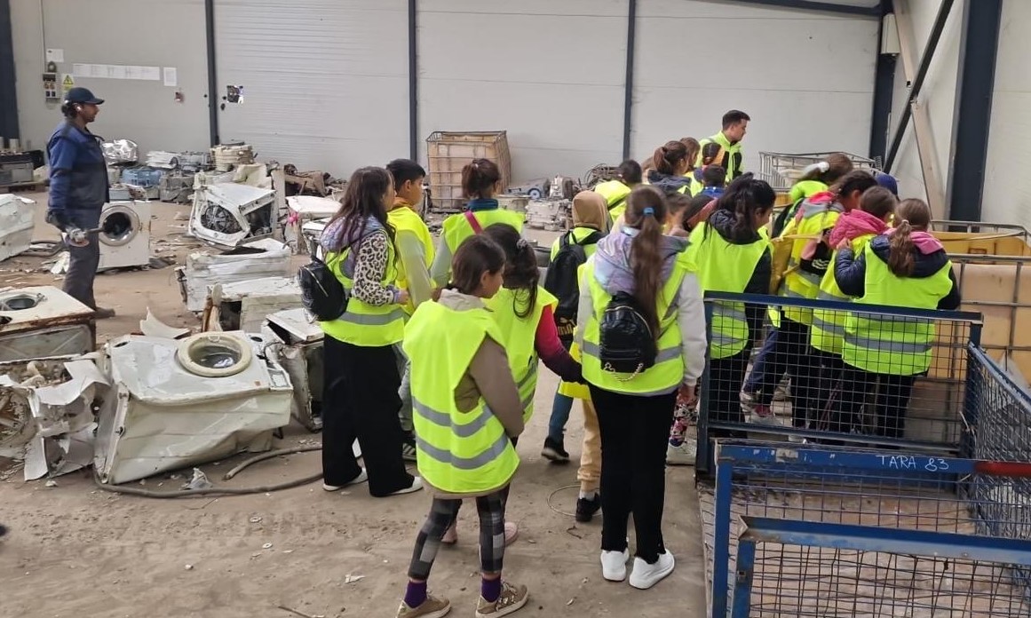 A group of students wearing high-visibility vests on an educational visit at a recycling facility, observing dismantled appliances and electronic waste, with a facility worker looking on.