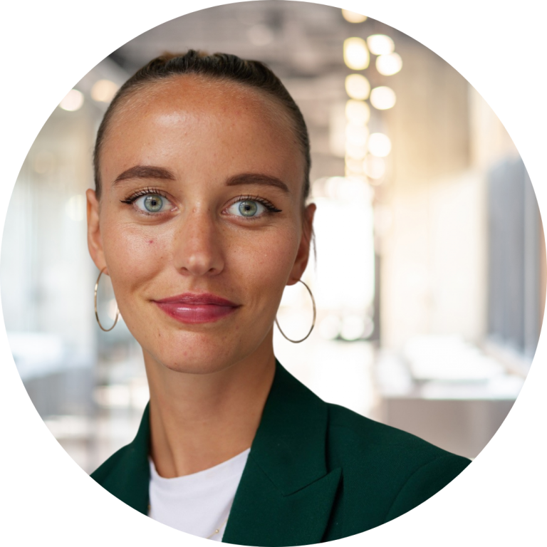 Close-up circular portrait of a young woman with green eyes, freckles, and slicked-back hair, wearing hoop earrings and a dark green blazer over a white top, smiling slightly against a softly lit, blurred office background.