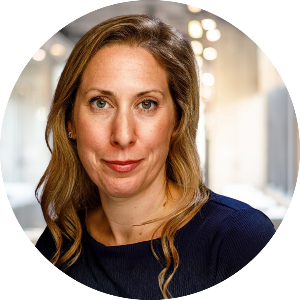 Head-and-shoulders portrait of a woman with light brown wavy hair, green eyes, and a slight smile, wearing a dark navy top against a softly lit modern interior background.