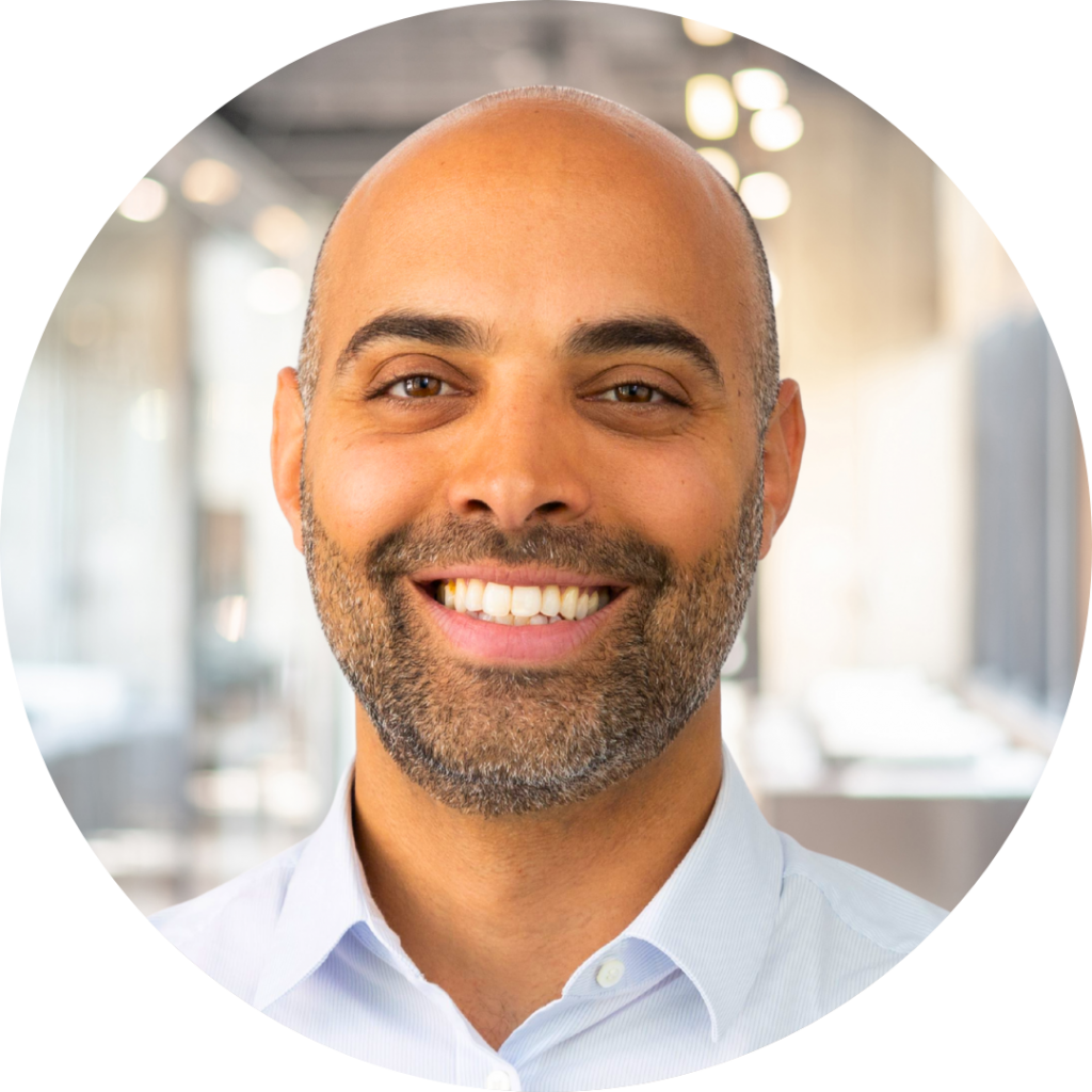 Smiling bald man with a trimmed salt-and-pepper beard wearing a light blue button-down shirt, close-up portrait in a bright, blurred modern office setting.