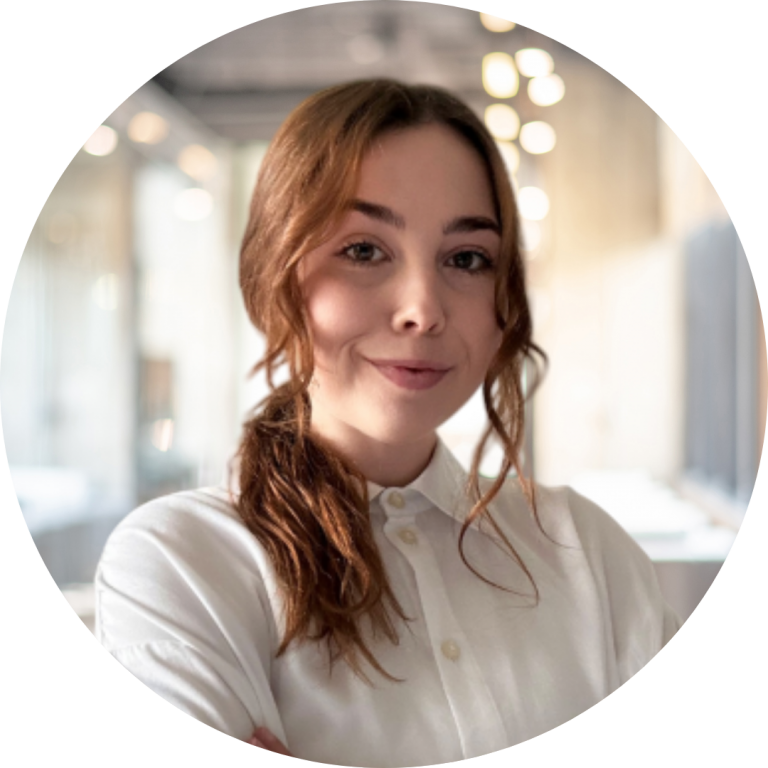 Young woman with reddish-brown hair in a loose ponytail wearing a white button-up shirt, smiling slightly with arms crossed in a softly lit modern office background.
